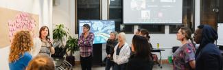 A group of team members gathers around a presenter who is speaking and pointing at a wall covered in pink sticky notes, engaging attentively in the session. In the background, others participate via a video call displayed on a large screen.