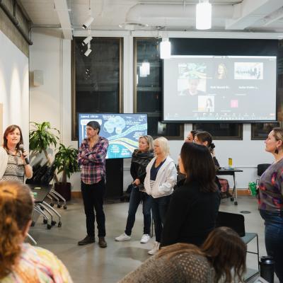 A group of team members gathers around a presenter who is speaking and pointing at a wall covered in pink sticky notes, engaging attentively in the session. In the background, others participate via a video call displayed on a large screen.